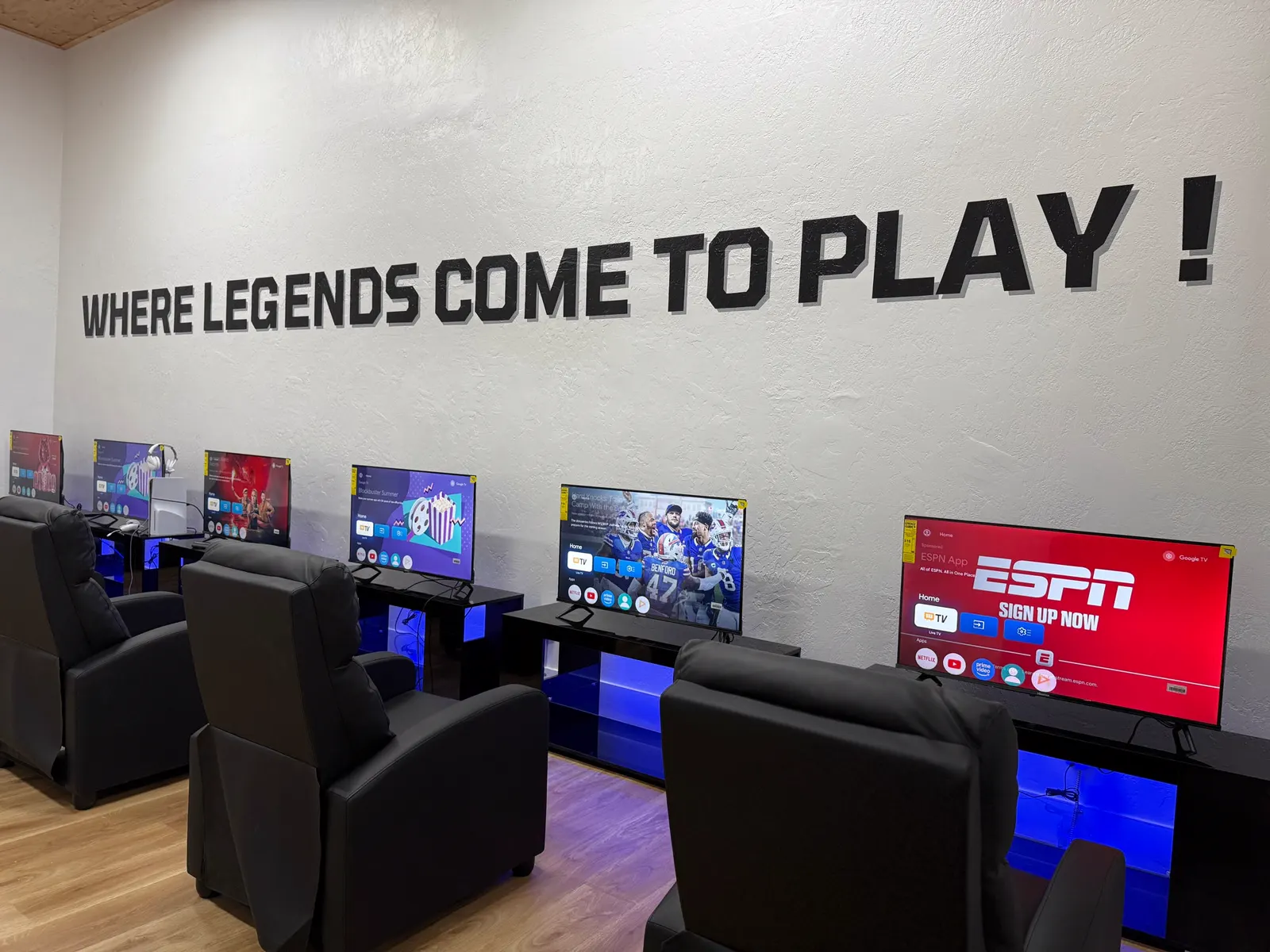 A row of gaming recliners facing TV stations under the wall lettering Where Legends Come to Play